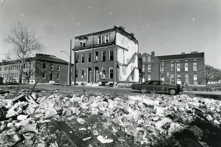 At the corner of Mills and N. Leffingwell a deteriorated, abandoned building sits across from a pile of debris and junk, 1973
