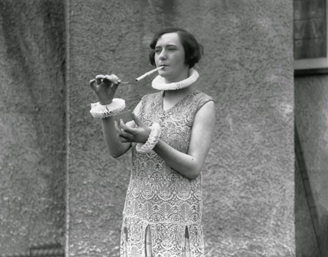 A woman smoking a large cigarette and wearing ruffles at collar and cuff, 1926.