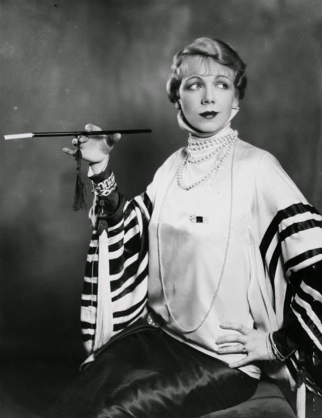 Actress Marjorie Brooks smoking a cigarette in a long cigarette holder in a production of The High Road, 1927.