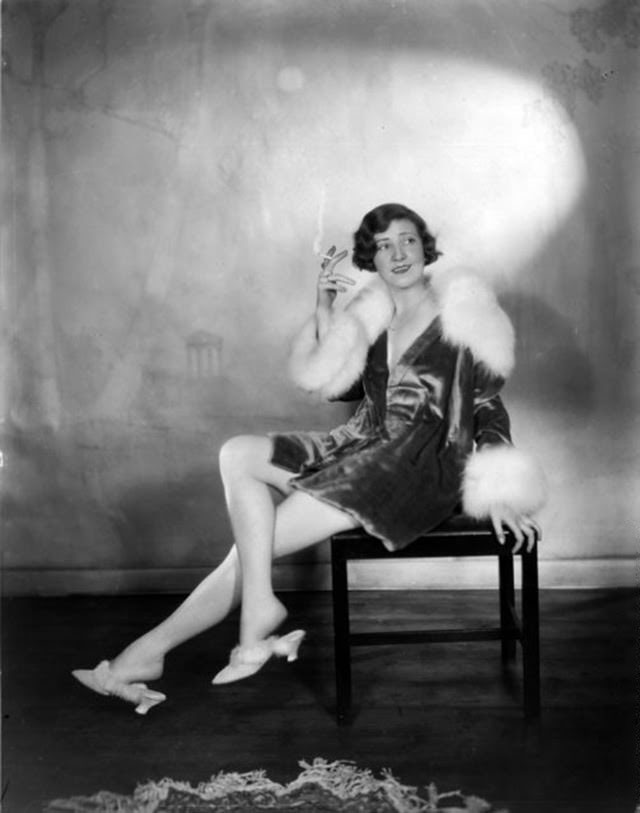 Actress Margaret Campbell, wearing a fur-trimmed outfit with matching slippers, smoking a cigarette, 1928.