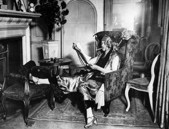 A lady wearing a smoking suit, reading a newspaper by the fireplace, 1922.