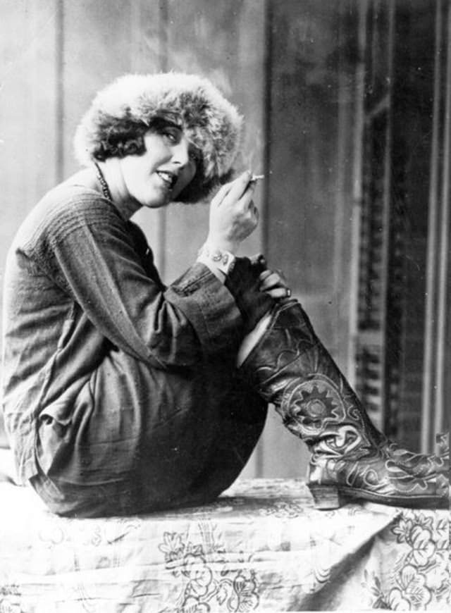 American journalist Louise Bryant poses for a portrait while wearing a Russian costume with decorated Western cowboy boots, sitting with her knees drawn up, smoking a cigarette, 1920