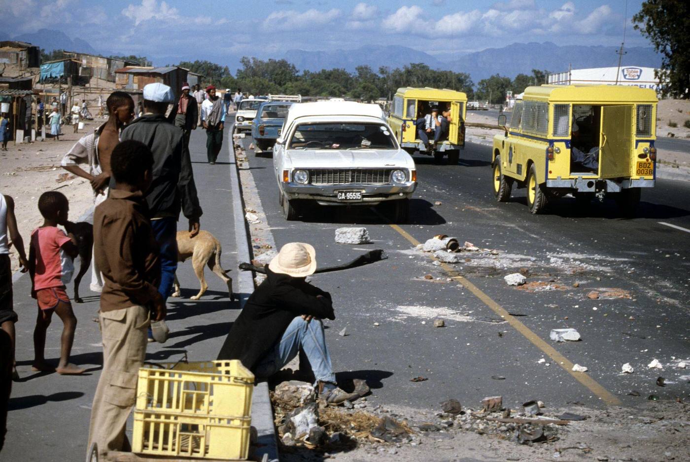 People on a sidewalk watch as South Africa Police Land Rovers drive past debris on a road in the Crossroads shantytown, Cape Town, 1985