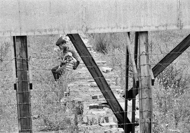A young boy playing with an East German border guard near a barbed wire fence along the border between East and West Berlin. The boy is looking through binoculars into the West.