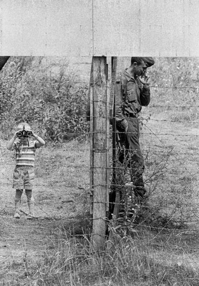 A young boy playing with an East German border guard near a barbed wire fence along the border between East and West Berlin. The boy is looking through binoculars into the West.