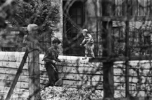 A boy playing with an East German border guard behind a barbed wire fence along the border wall between East and West Berlin.