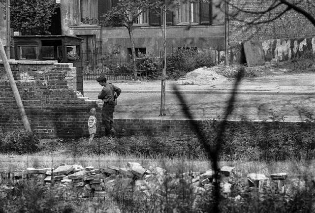 A boy playing with an East German border guard behind a barbed wire fence along the border wall between East and West Berlin.