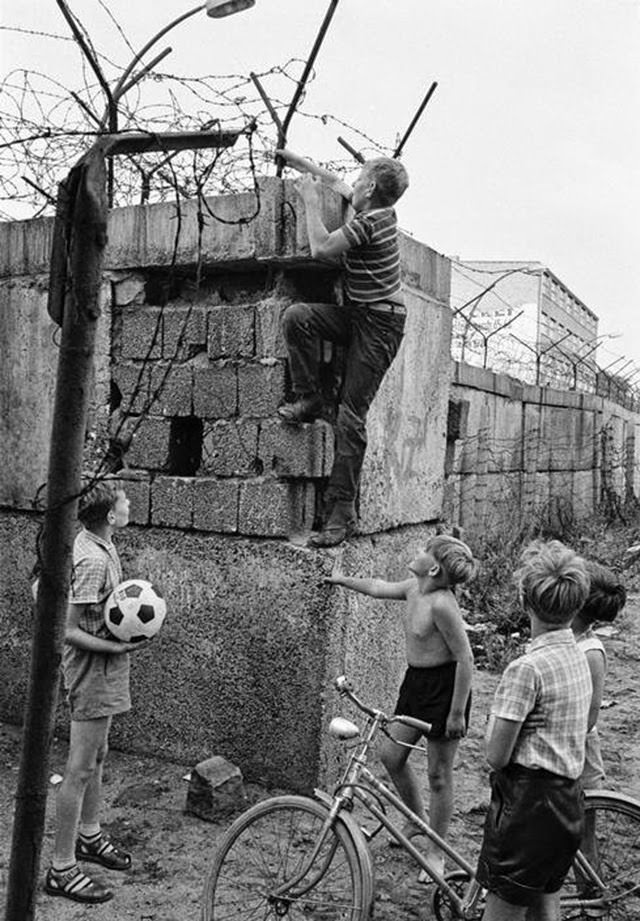 Scene at the Berlin Wall near Bernauer Strasse. Four children, one of them with a soccer ball and another with a bike, watching a fifth child climbing up the Wall.