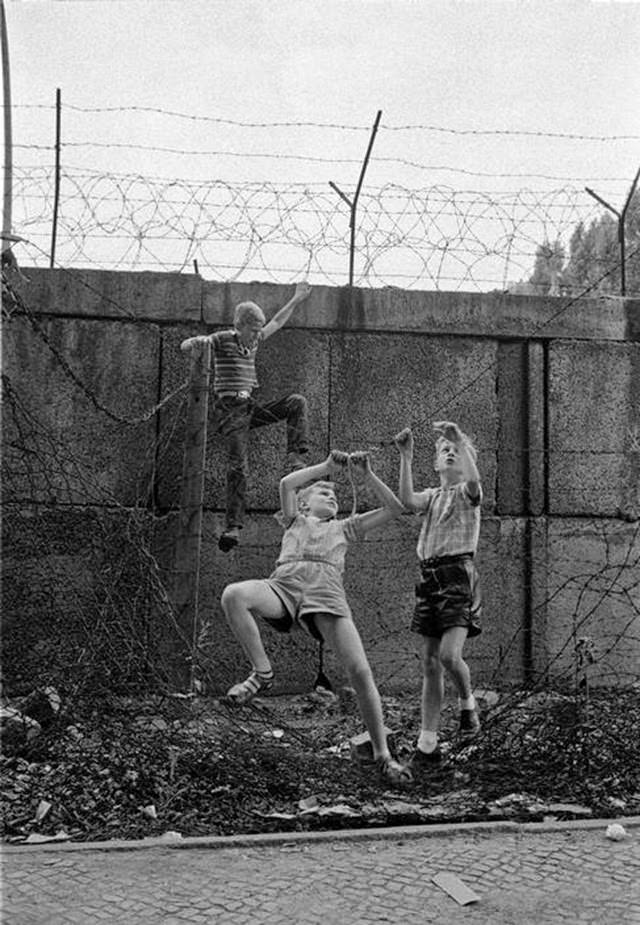 Children playing at the Berlin Wall near Bernauer Strasse. The three boys are climbing up the wall and moving along a barbed wire fence alongside the wall hand by hand.