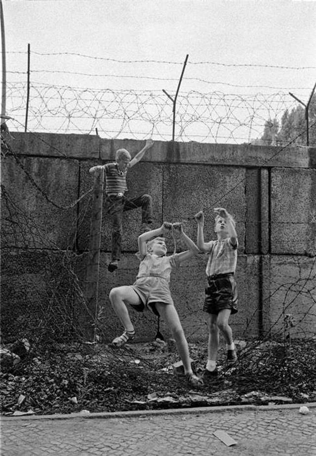 Children playing at the Berlin Wall near Bernauer Strasse. The three boys are climbing up the wall and moving along a barbed wire fence alongside the wall hand by hand.