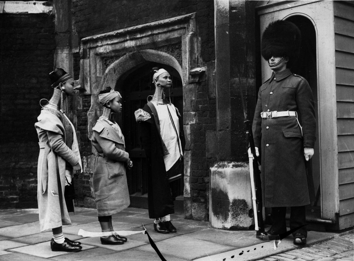 Giraffe necked women from Burma looking at a soldier standing guard in London, 1935.