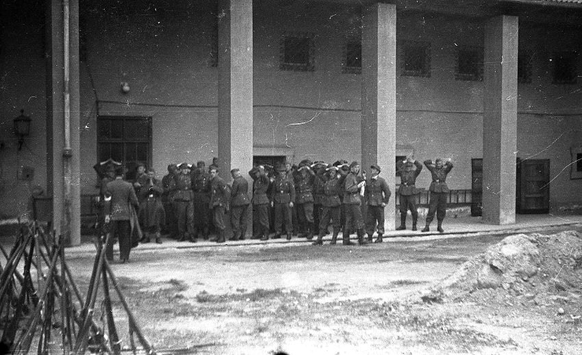 The men in the background here have their hands raised. They are possibly the German camp guards who surrendered and turned in their weapons, seen in the left foreground of the picture.