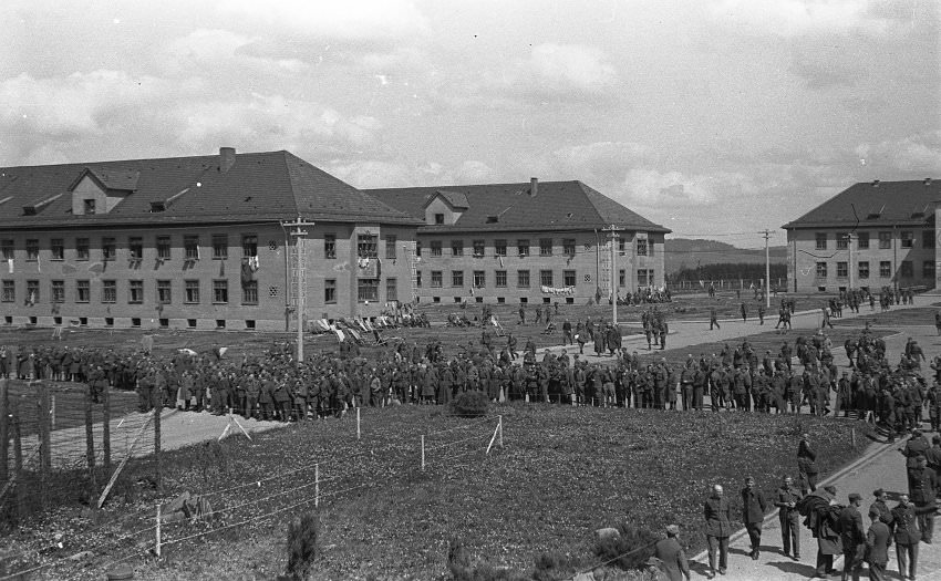 A relaxed atmosphere pervaded in the camp after its liberation by American forces in 1945. In front of the barracks on the left, some former inmates sit on lounge chairs in the sun.