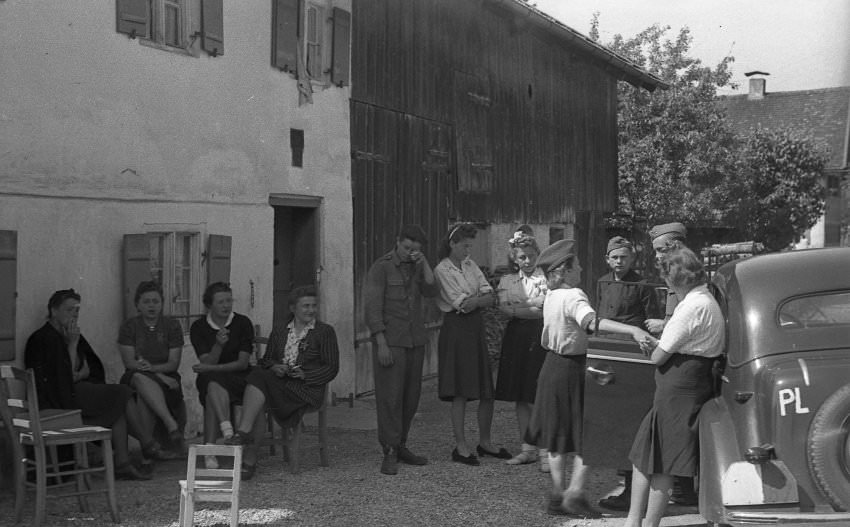 A few uniformed men stand relaxed by a car and converse with some women. Other women in civilian clothes sit by and smoke. Who are these people and what led the photographer to capture them in film?