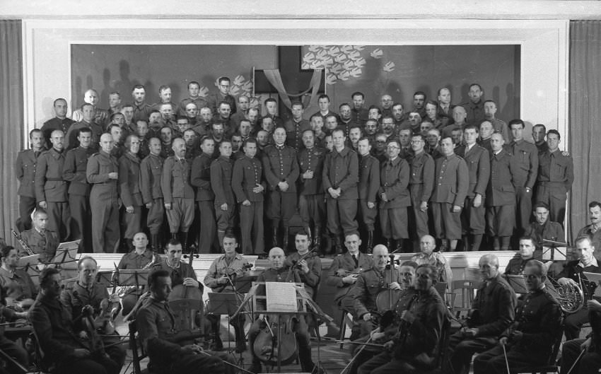 A group of officers poses on the stage of the camp theater, with the orchestra in the foreground.
