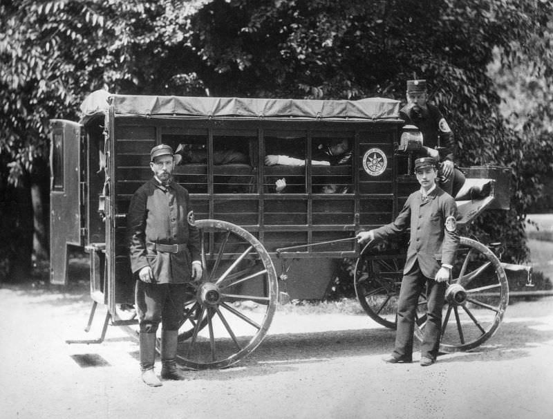 A Wiener Ambulance with patients in ‘layers’ in a horse drawn wooden carriage. The sides are partly open, but have curtains. The ambulance men are members of the Viennese Voluntary Rescue Society founded in 1881.