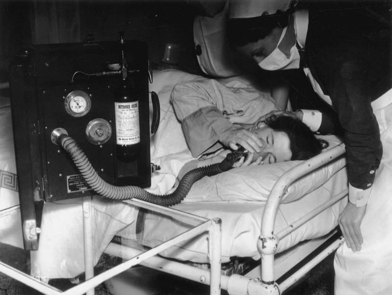 In an effort to make childbirth as painless as possible, a patient inhales analgesia during labor whilst a nurse looks over her, July 1939.