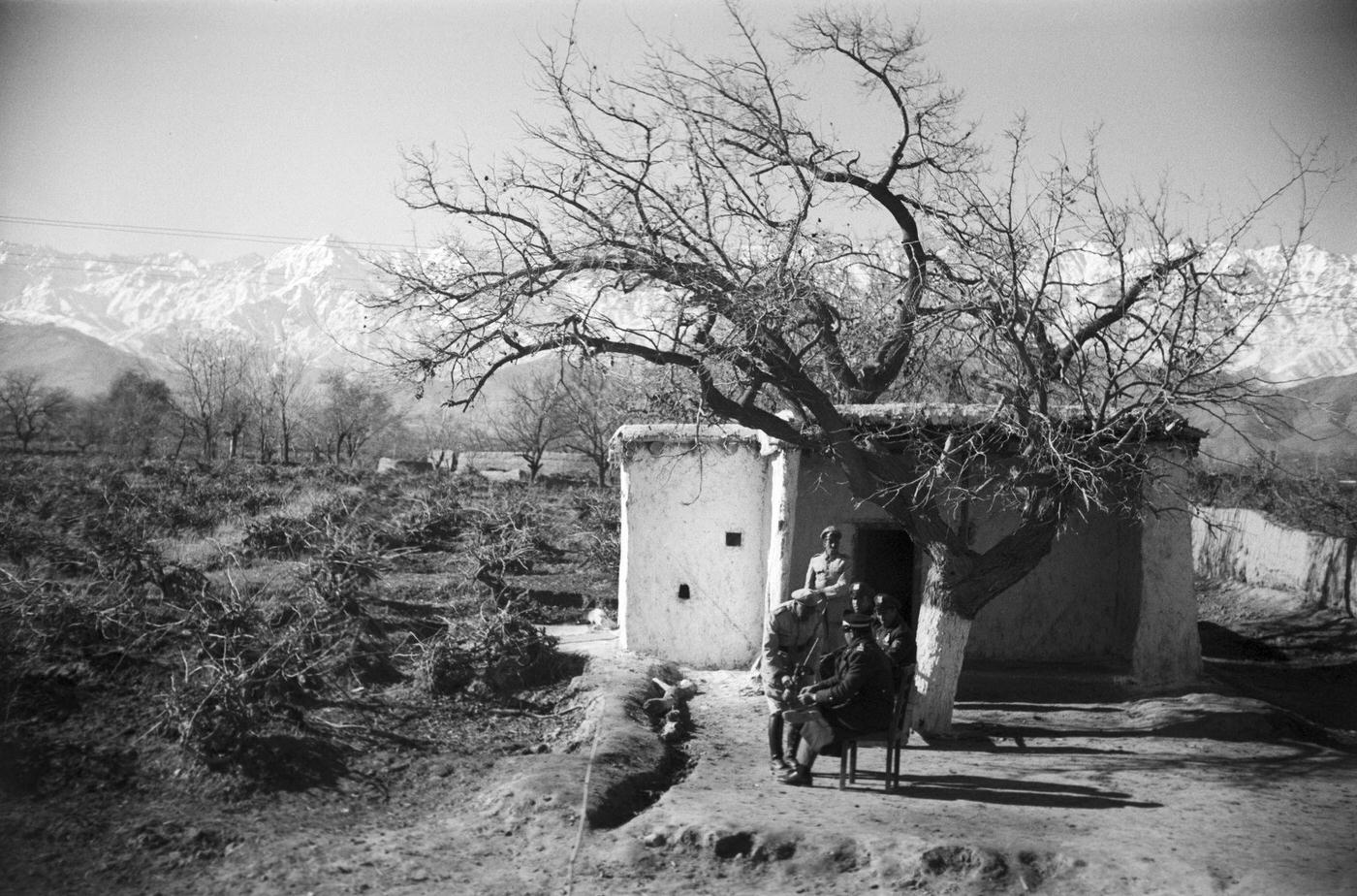 Soldiers, seated near a tree, in front of a dirt building, on the road to Kabul