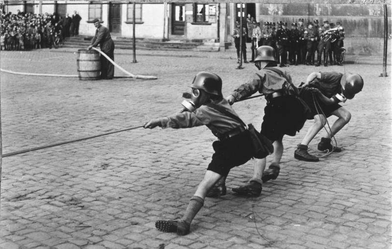 Hitler Youth members playing tug of war while donning helmets and gas masks, 1933.