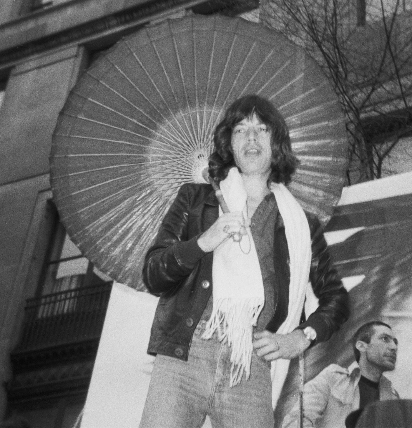 Mick Jagger poses with a parasol, while Charlie Watts is visible, as The Rolling Stones perform on a flatbed truck on Fifth Avenue, New York City, 1975.