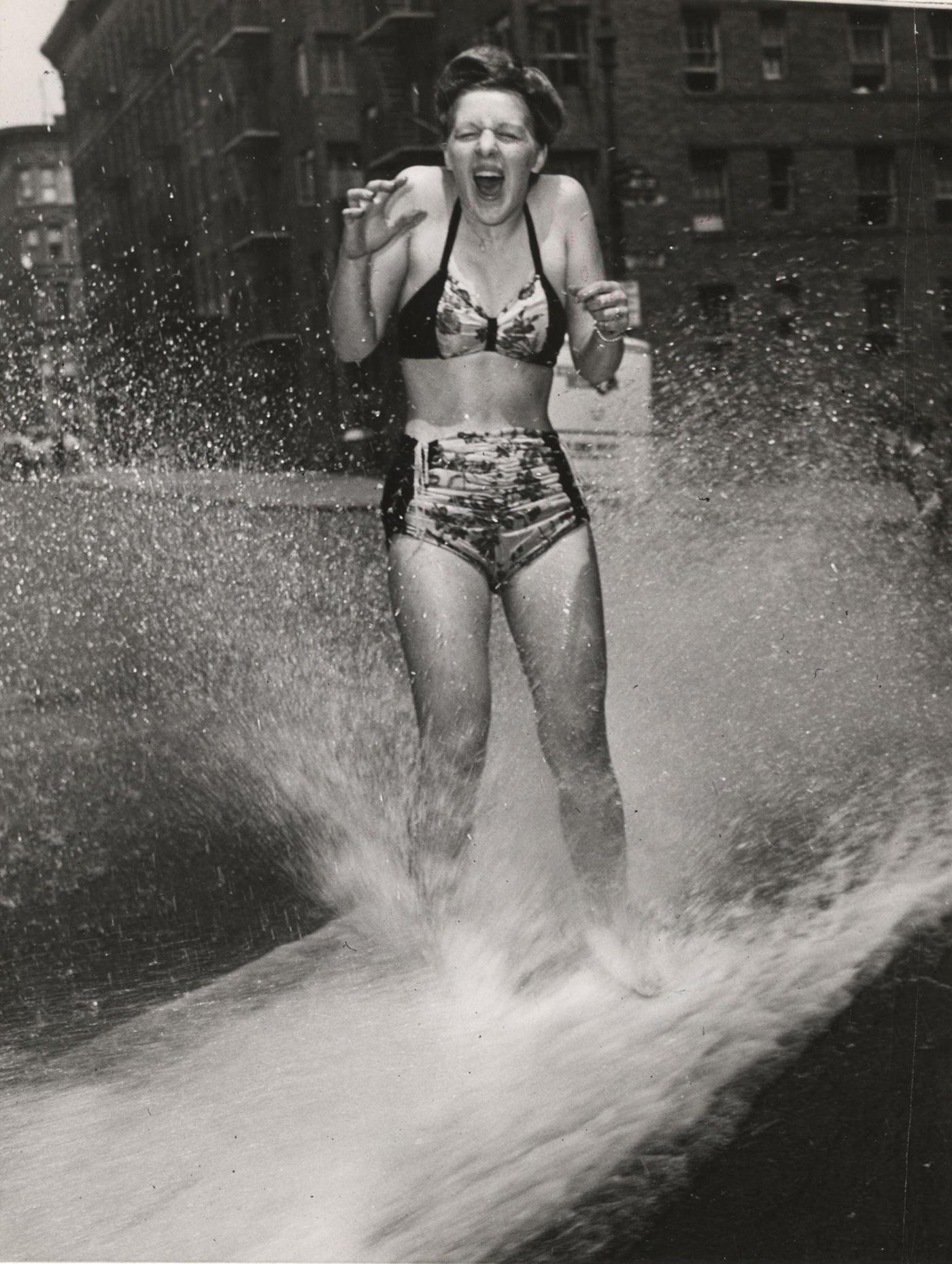 Woman in a two-piece bathing suit cools down under a fire hydrant spray, 1930s