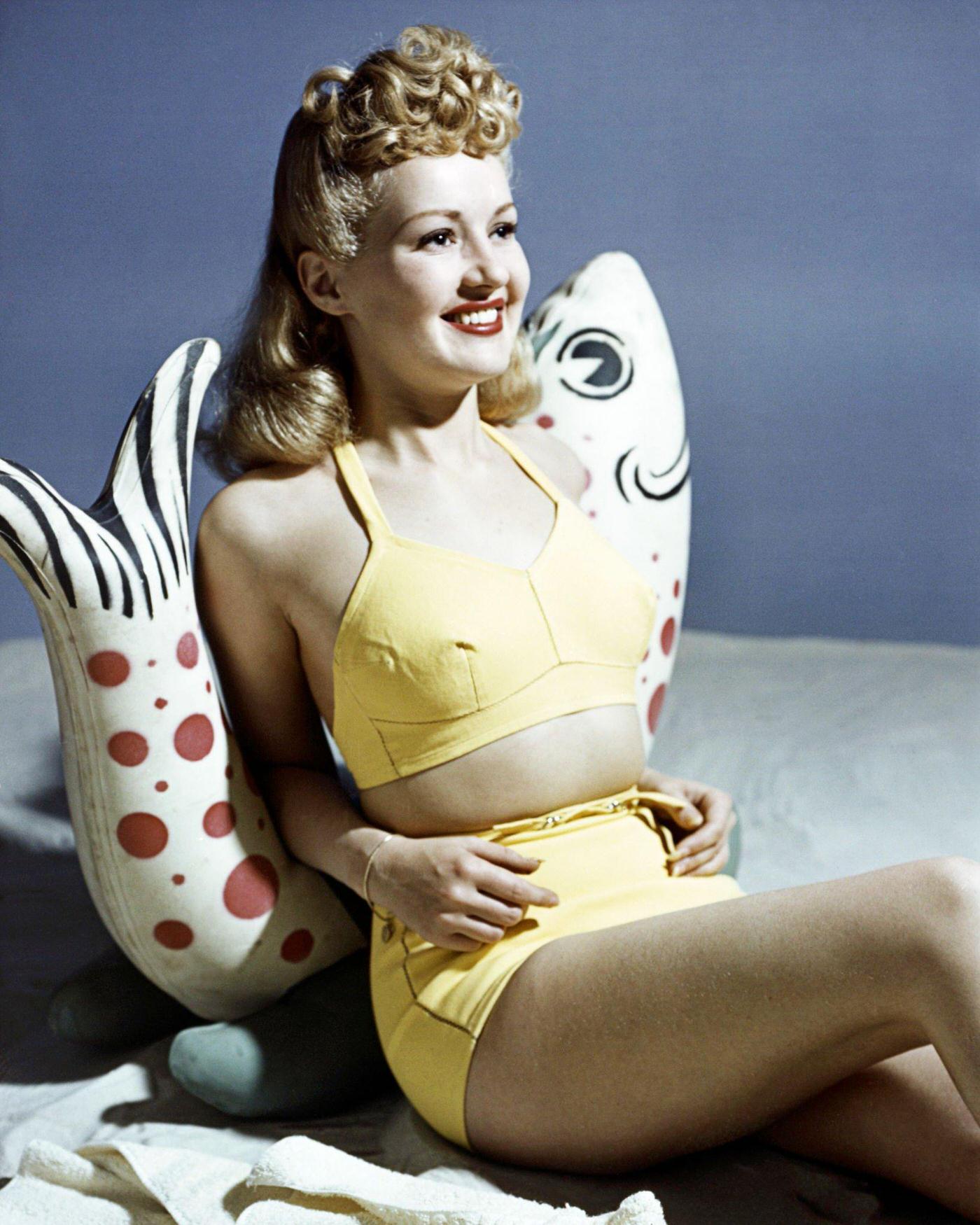 Betty Grable, US actress, in a yellow bikini leaning against a fish ornament, 1940.