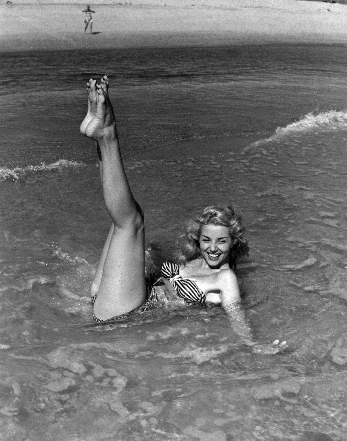 Young woman laying on the seashore wearing a striped bikini, 1948.