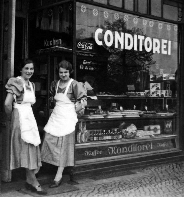 Two ladies pose in front of a Konditorei in Germany displaying a Coca-Cola sign, circa WWII