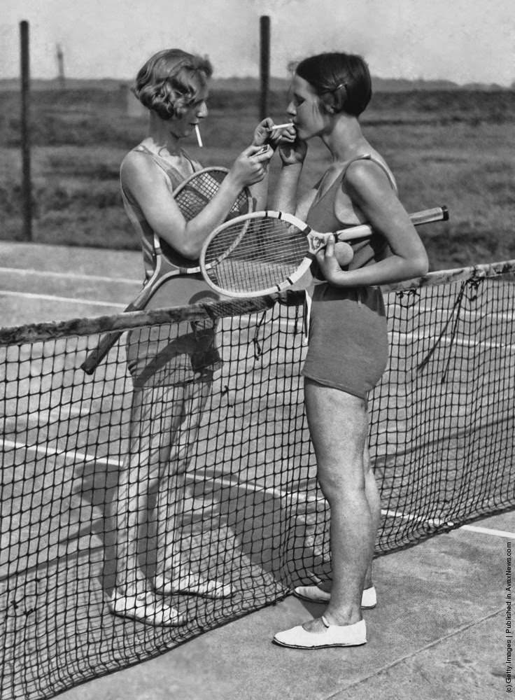 Two women lighting cigarettes on a tennis court in Essex, England 1930s.