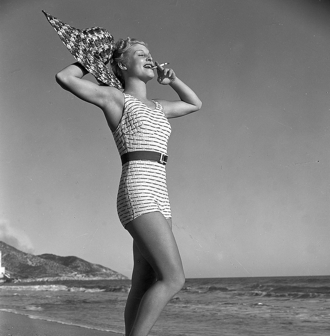 Blonde beauty in her bathing suit on the beach smoking a cigarette, 1930.