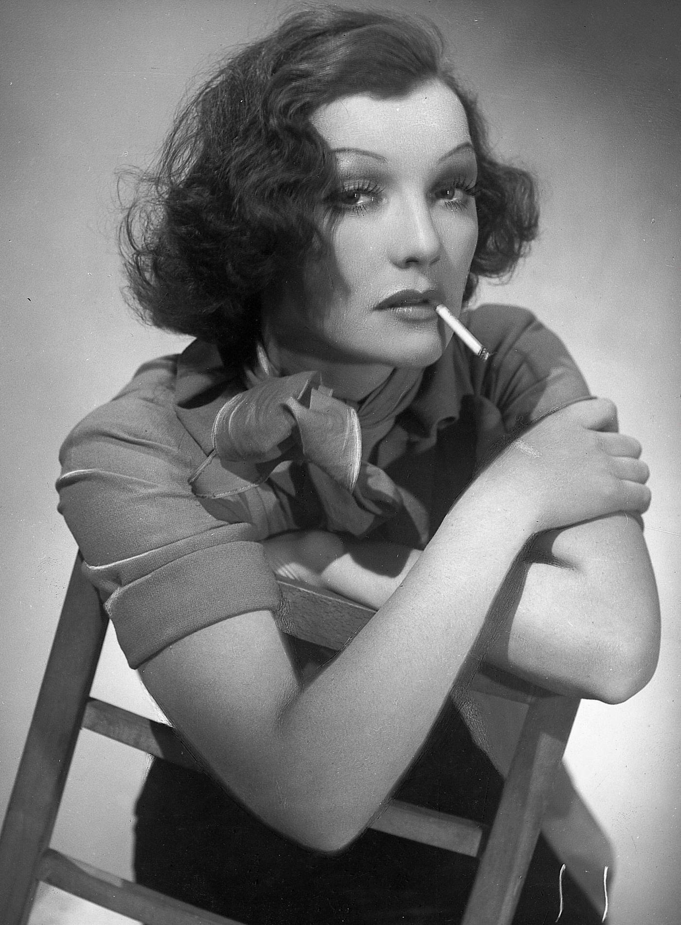 Brunette woman, rouged strongly, sits on a chair and smokes a cigarette, 1930.