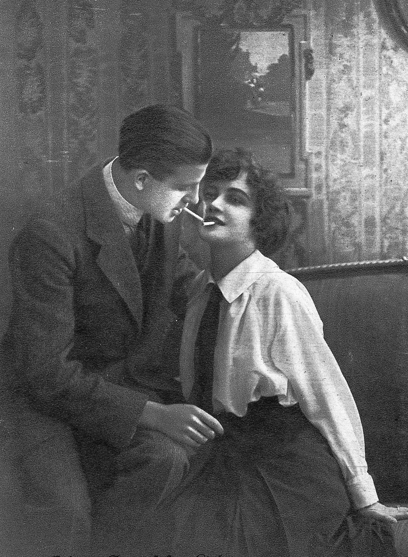 Young man bends over to a woman sitting on the sofa, lights his cigarette with the glow of hers, 1930.