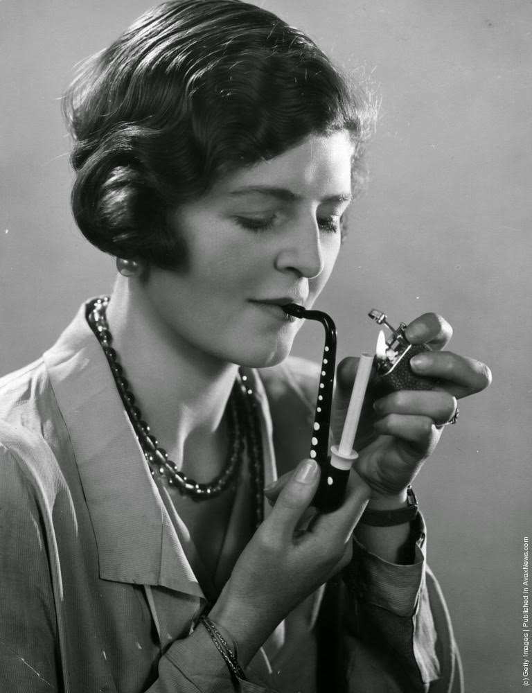 A lady is pictured lighting her cigarette now held in a novelty cigarette holder in the shape of a musical instrument, 1932
