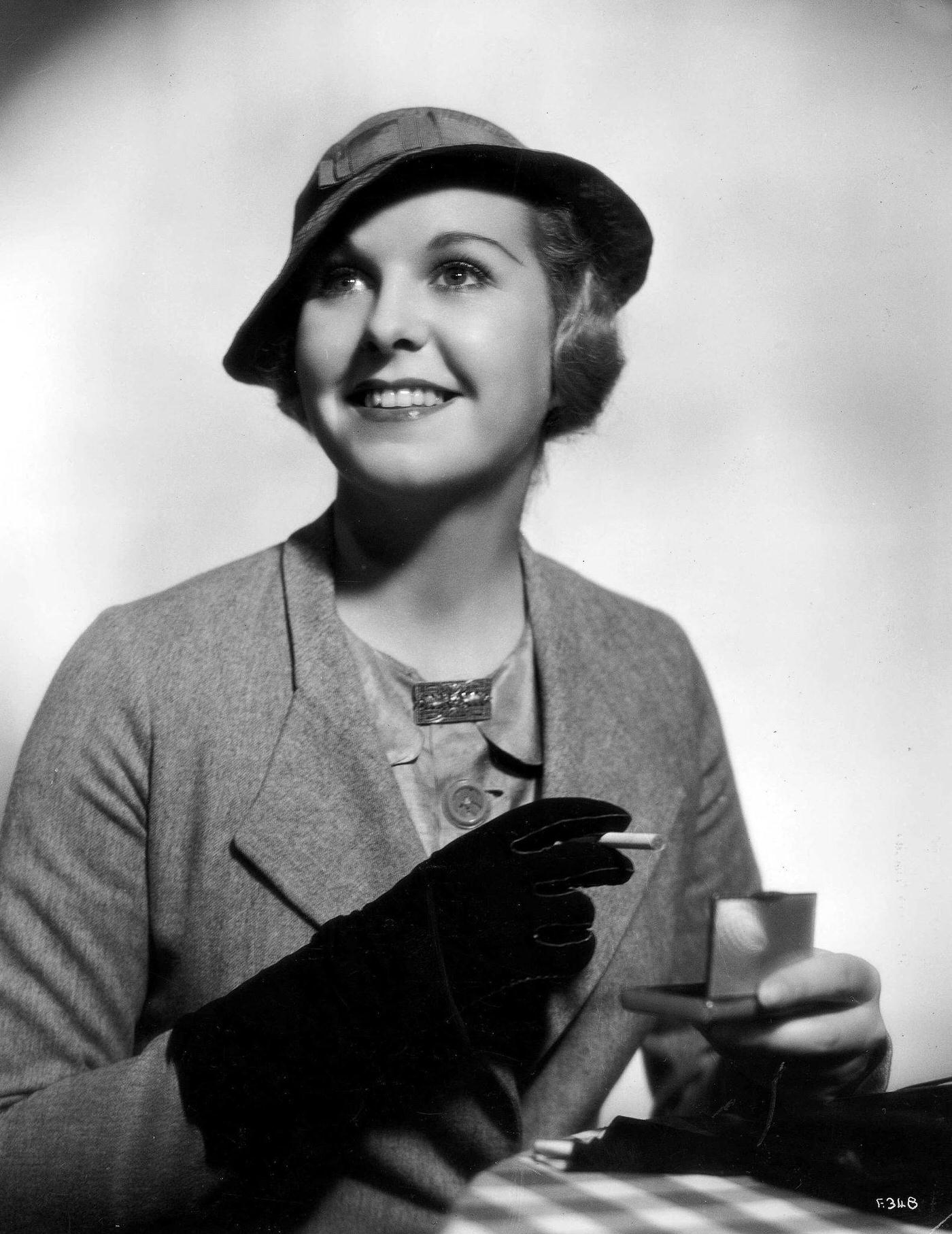 A lady wearing a grey flannel suit with a stitched hat and black gloves about to light up a cigarette, 1935