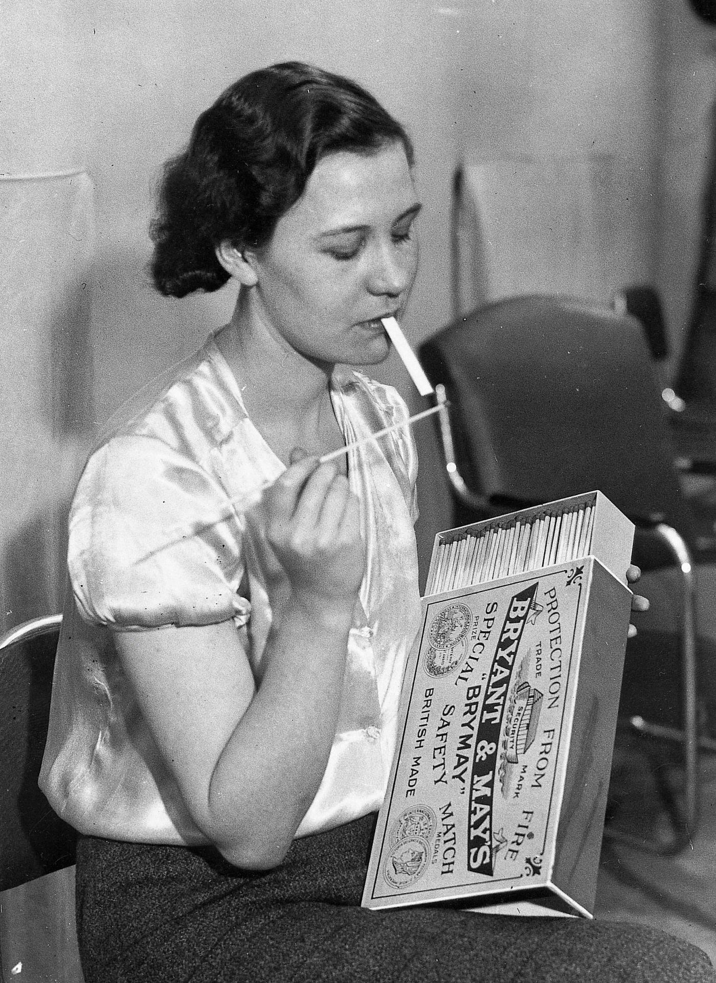 A woman is lighting a cigarette with oversized matches at the 39th International Tabacco Exhibition, London, 1935