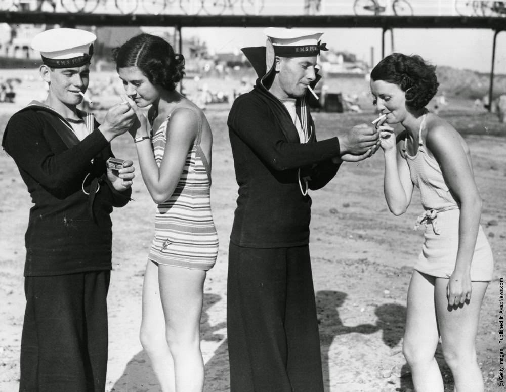 Two sailors from HMS Fury enjoy a smoke with two women in bathing suits on a beach in Jersey, where their ship is on a visit, 1935