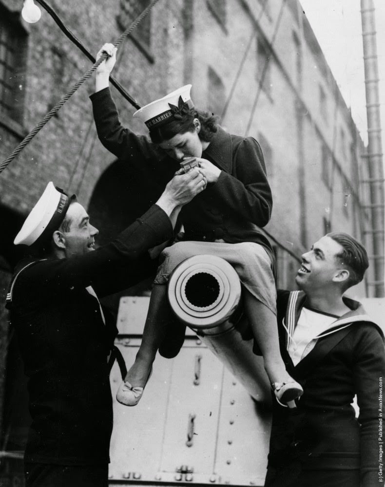 A young female British Navy officer sitting astride a minesweepers cannon and lighting a cigarette whilst two officers look on, 5th June 1937