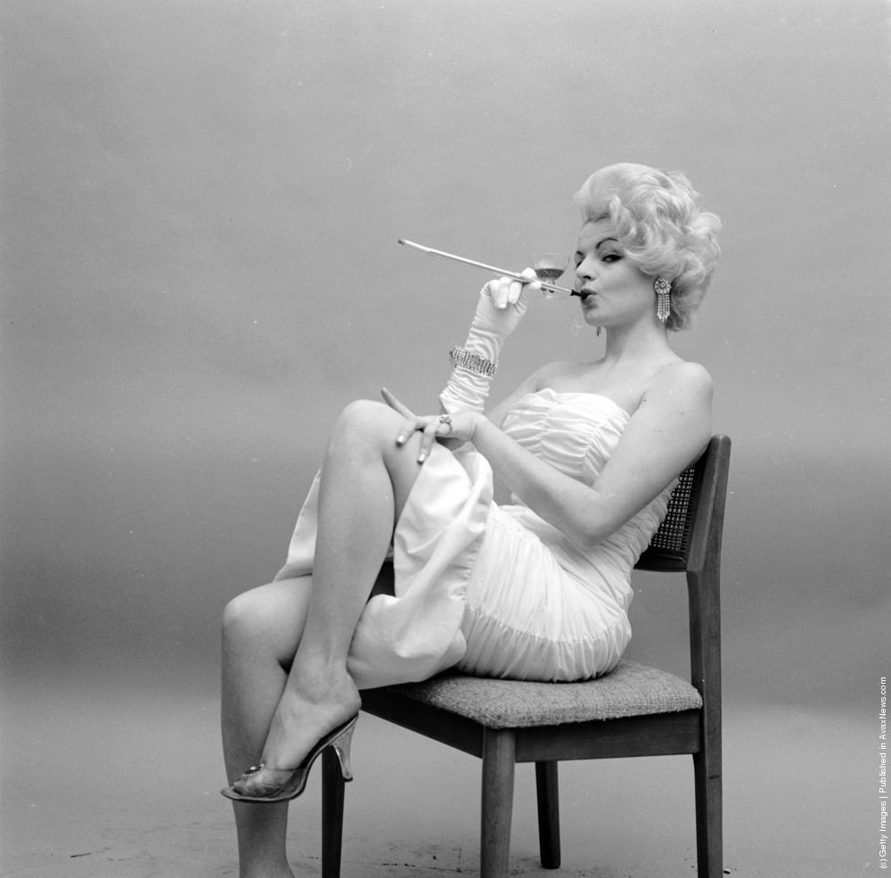 Model Bonnie Sharie uses a long cigarette holder with an unusual wine glass attachment, circa 1955.