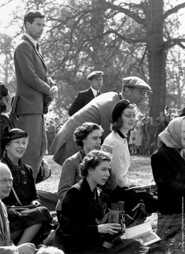 Queen Elizabeth II holding a camera at the Olympic Horse Trials at Badminton, whilst Princess Margaret sits behind her, smoking a cigarette and watching the action. Also in the group is Group Captain Peter Townsend (left). 13th October 1955.