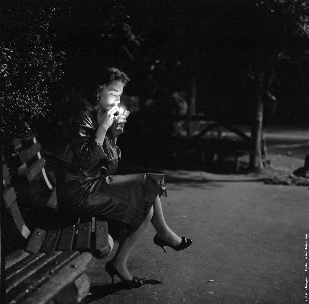 A young woman lighting a cigarette as she sits on a New York park bench at night, circa 1957.