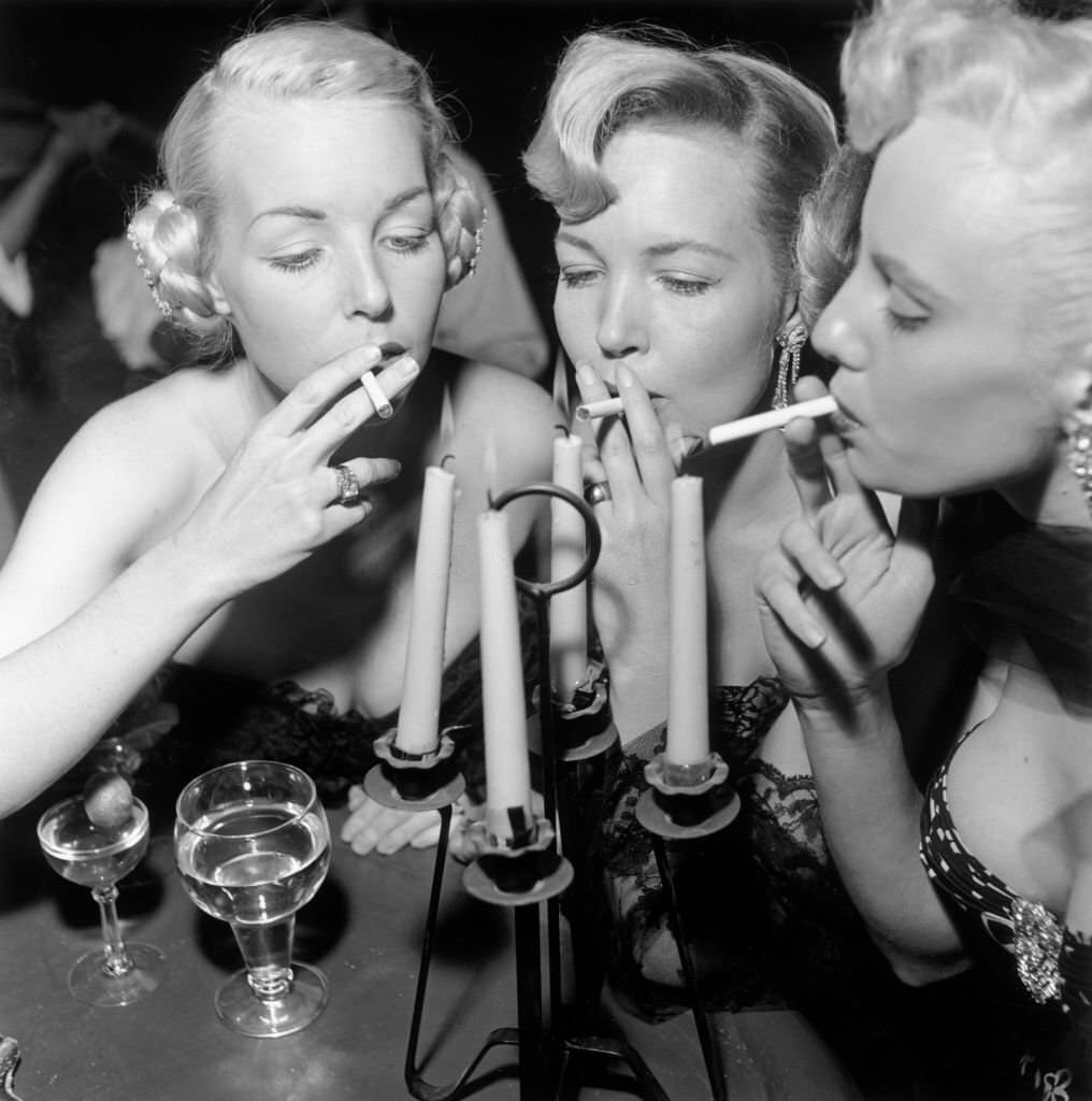 Three aspiring actors Shirley Buchanan, Mara Lynn, and Donna Williams light their cigarettes from a candelabra at a bar, Loreto, Mexico, 1950s.