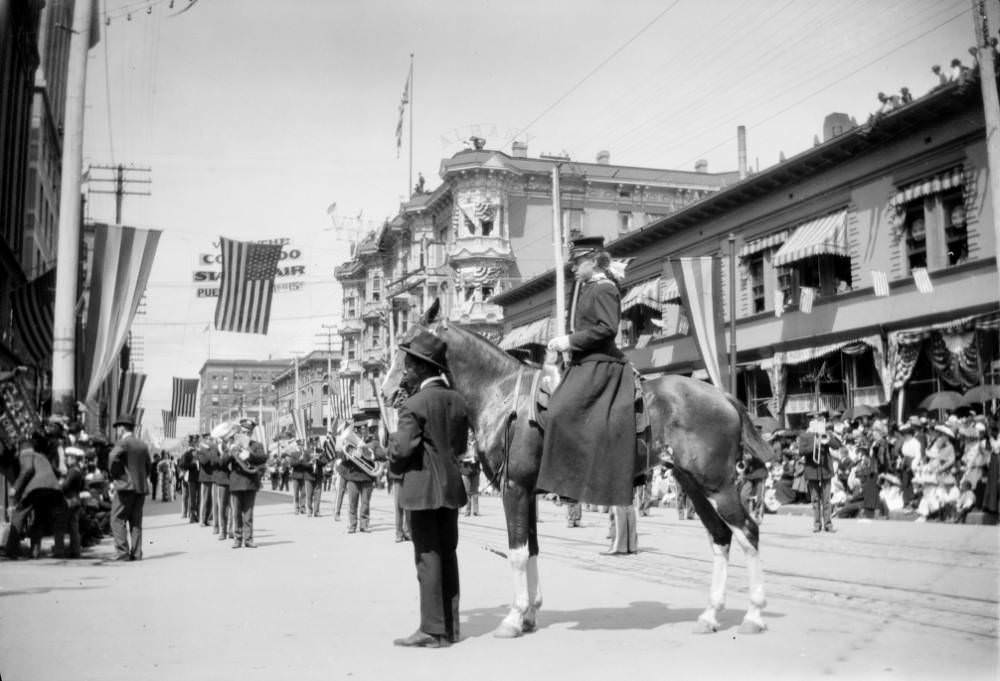 African American man and woman on horseback participate in a parade on 17th Street, Denver, Colorado, marching band and U.S. flags visible, 1900s