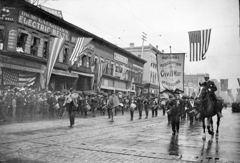 Parade on 17th Street, Denver with marching band, spectators, and signs on buildings for various businesses, 1900s
