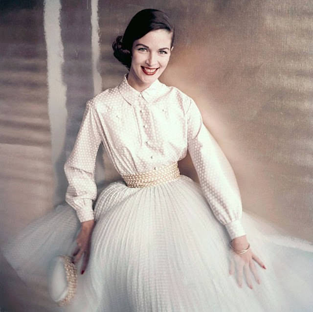 Theo Graham wears a pale pink shirt and pleated blue skirt with white dots, accented by a yellow dotted cummerbund, 1955.