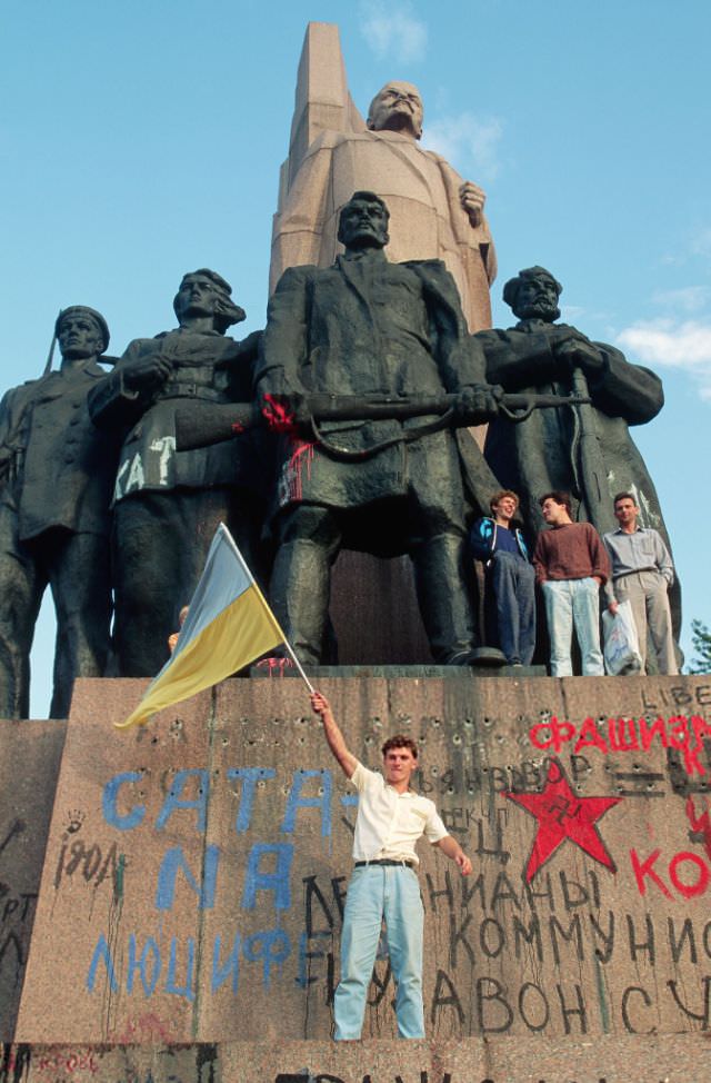 A young man waves the Ukrainian flag on the heavily graffitied base of a statue of Lenin while others watch, Ukraine, 1991
