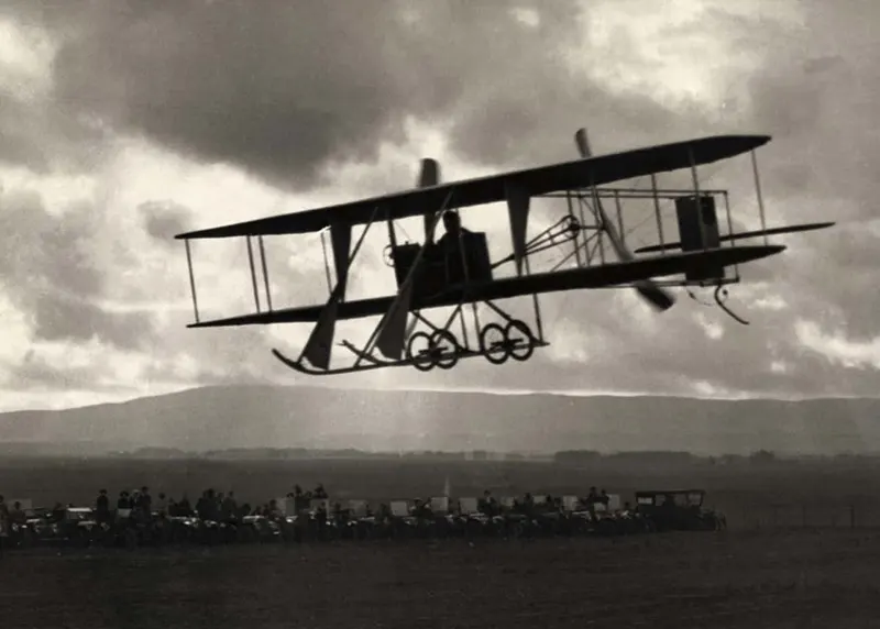A Wright Brothers plane flying by spectators.
