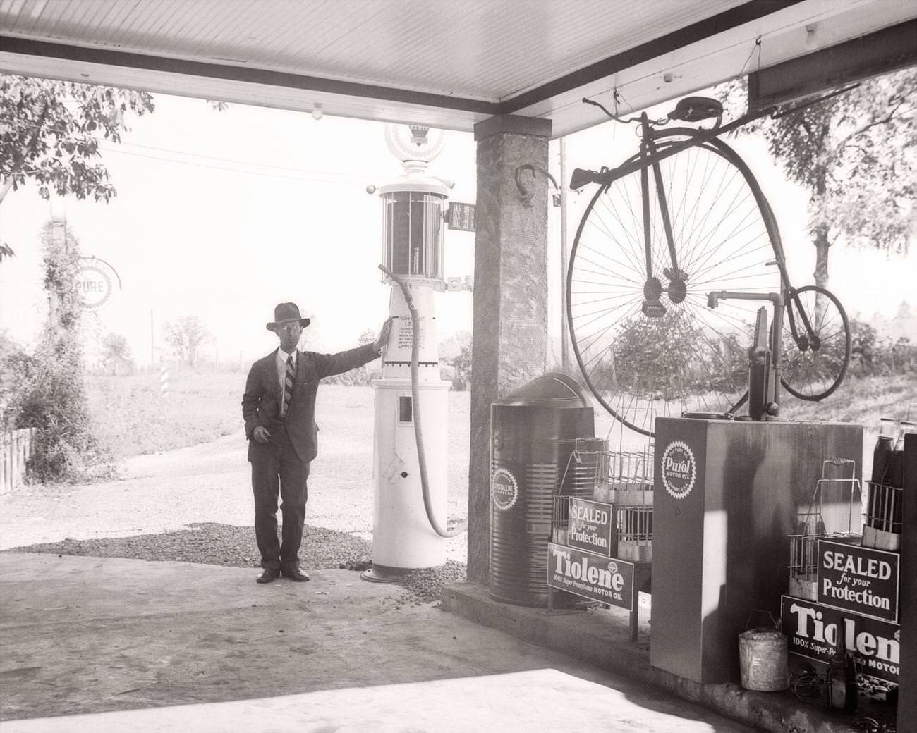 Man leaning on gas pump at filling station in Ohio with antique displays, 1940s.