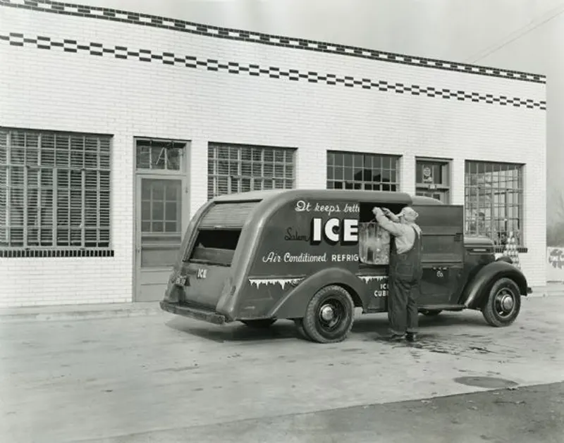 A man lifts a block of ice, using tongs, from an International D-15 truck owned by the Salem Ice Company as he makes a delivery to what appears to be a restaurant.
