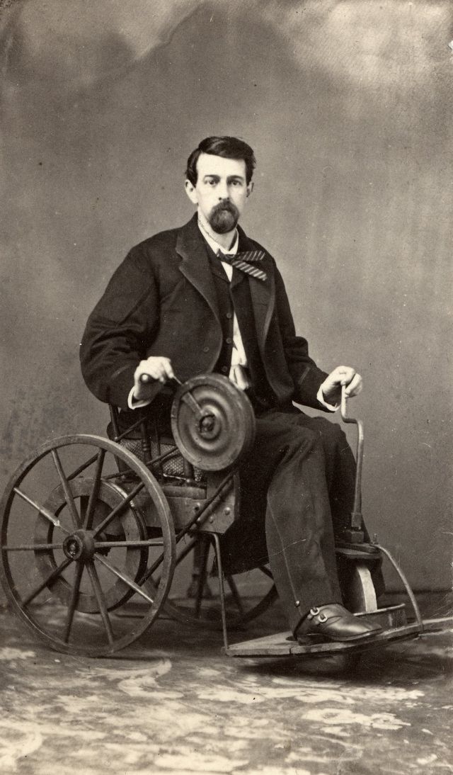 A man sits in a wheel chair that features a flywheel with hand-crank that turns a belt and powers the large back wheels