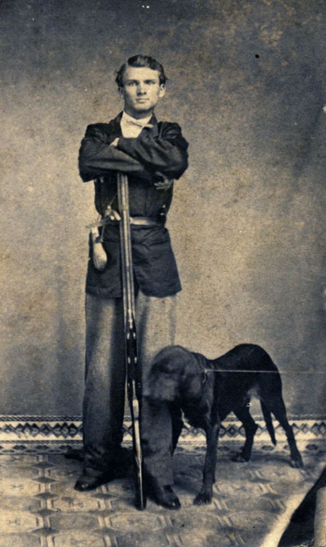 A thin young man, likely in his late teens or early twenties, stands with forearms resting atop the muzzle of his firearm and his dog